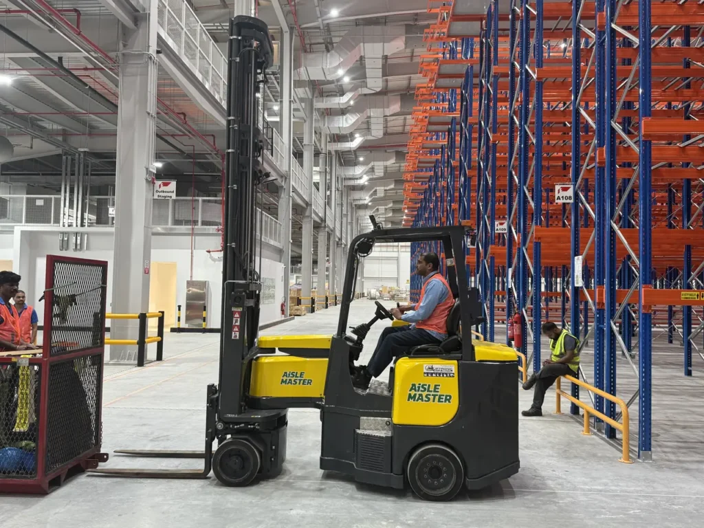 Operator driving an Aisle-Master articulated forklift past high-bay pallet racking