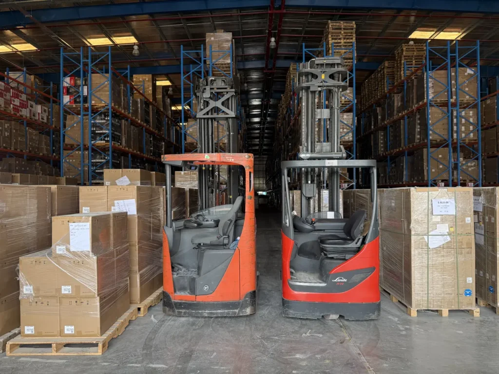 Two electric reach trucks between selective pallet racking and stacked cartons